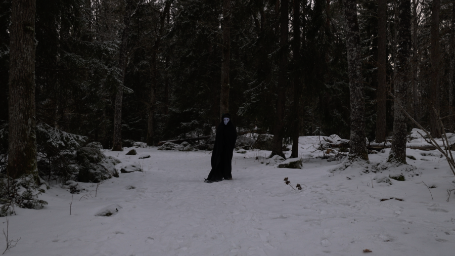 En m&ouml;rk skepnad med vit mask g&aring;r i en m&ouml;rk vinterskog.