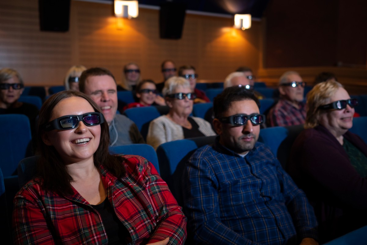 Bilden visar en grupp människor som sitter i en biosalong. De har på sig 3D-glasögon och verkar titta på en film. Personerna sitter i rader på blåa stolar. Kvinnan längst fram till vänster bär en röd- och svartmönstrad skjorta och ler. Det finns flera andra personer i bakgrunden, och belysningen i rummet är dämpad, vilket är typiskt för en biosalong.