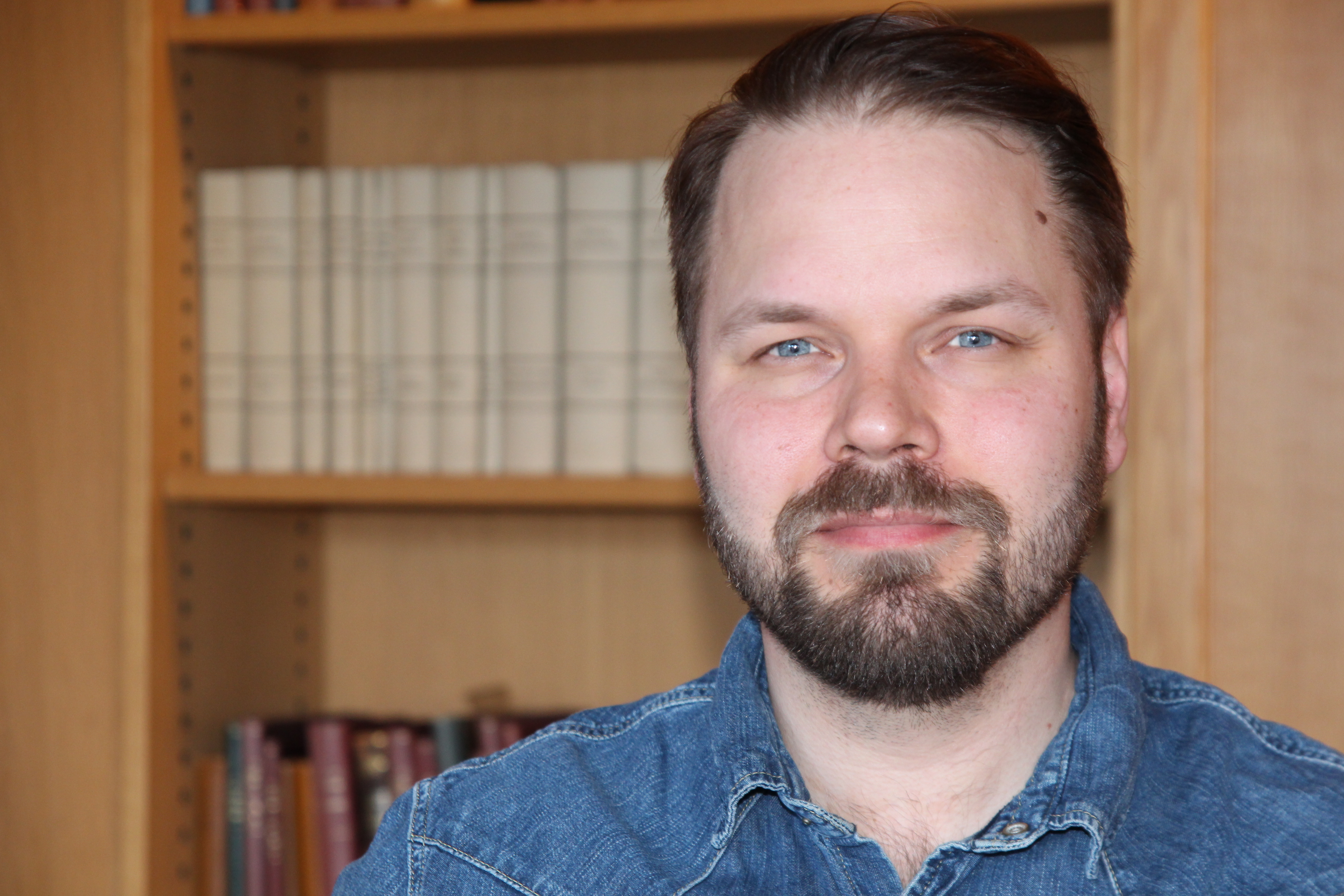 Markus Lindgren Miljöpartiet. Han är klädd i en mellanblå jeansskjorta med tryckknappar, har mörkt hår och skägg. I bakgrunden ser man en bokhylla fylld med gamla böcker i oskärpa.