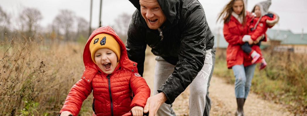 Familj utomhus med två barn och två vuxna, ett barn lär sig cykla.