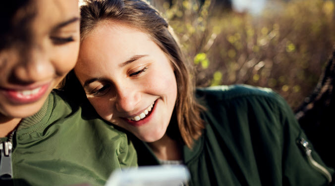 Två tjejer sitter ute och skrattar och läser på en mobil eller bok