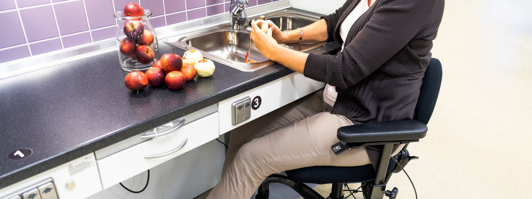 En kvinna sitter i en arbetsstol vid diskbänken och skalar äpplen