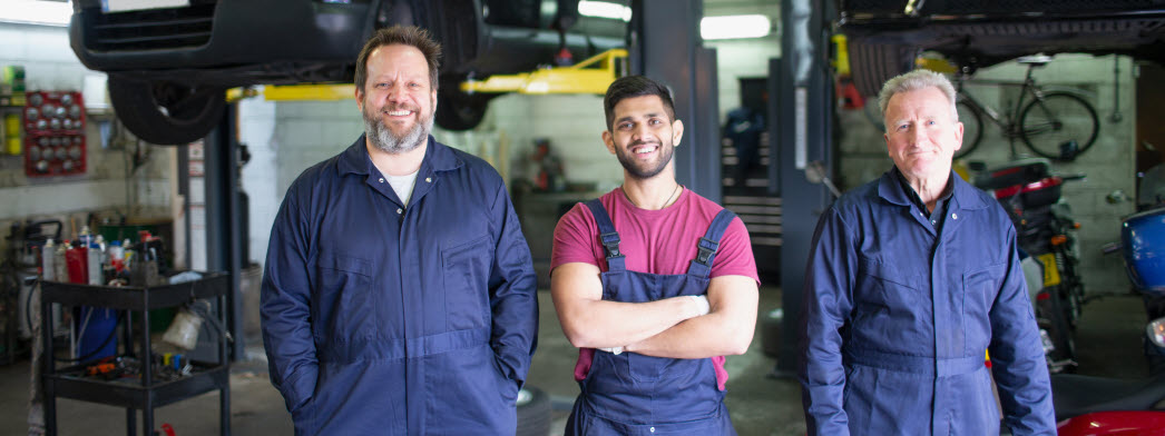 Tre män i olika åldrar och etniciteter klädda i blåa arbetskläder i en bilverkstad. Samtliga tittar framåt och ler.